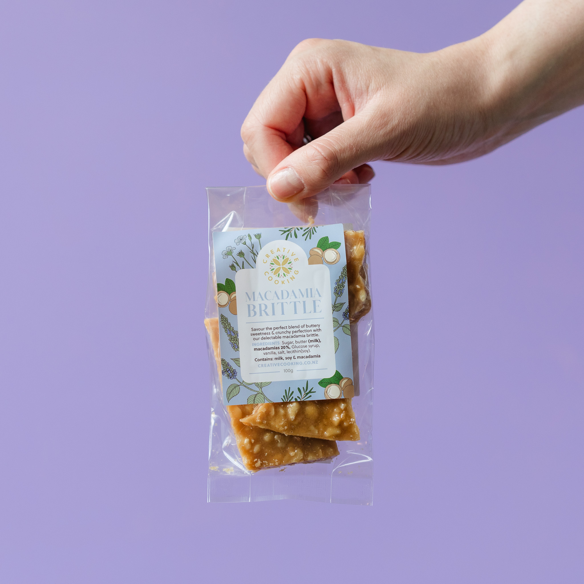 Macadamia Brittle by Superseed Crackers held by a hand against purple background